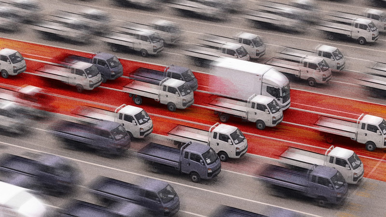 Vista aérea de una carretera muy transitada con efecto movimiento borroso, mostrando un atasco de tráfico de pequeños camiones y camionetas en diferentes tonos de gris y blanco, con un distintivo carril rojo en el centro.