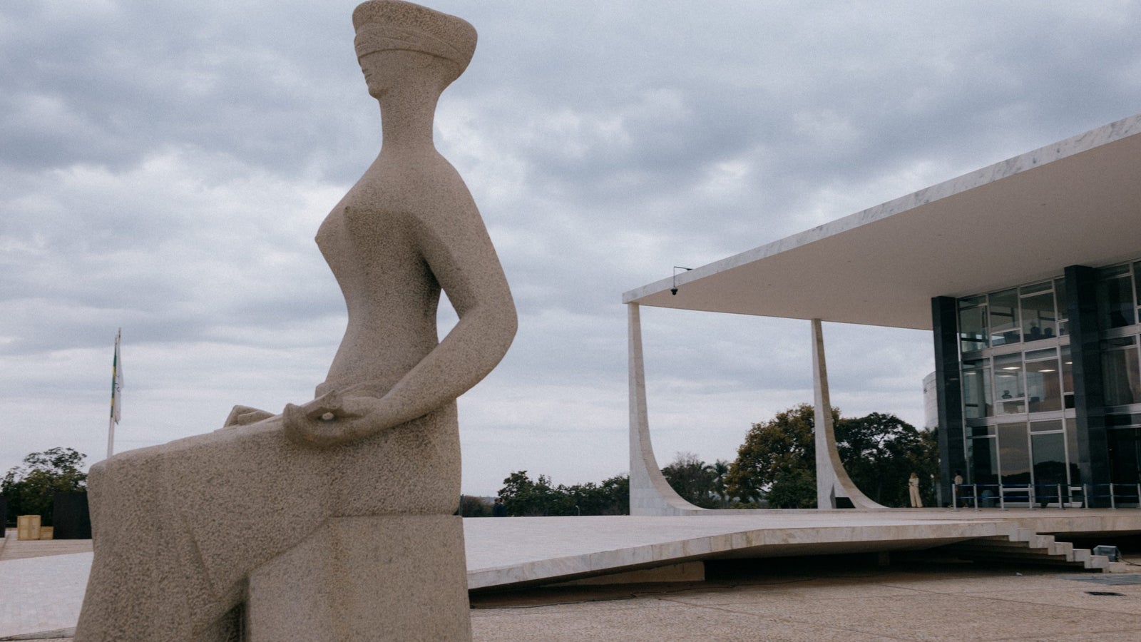 Una gran escultura de piedra de una mujer con los ojos vendados sentado con una expresión serena, situada en una plaza abierta frente a un moderno edificio arquitectónico con techo plano y grandes ventanales de cristal, bajo un cielo nublado.