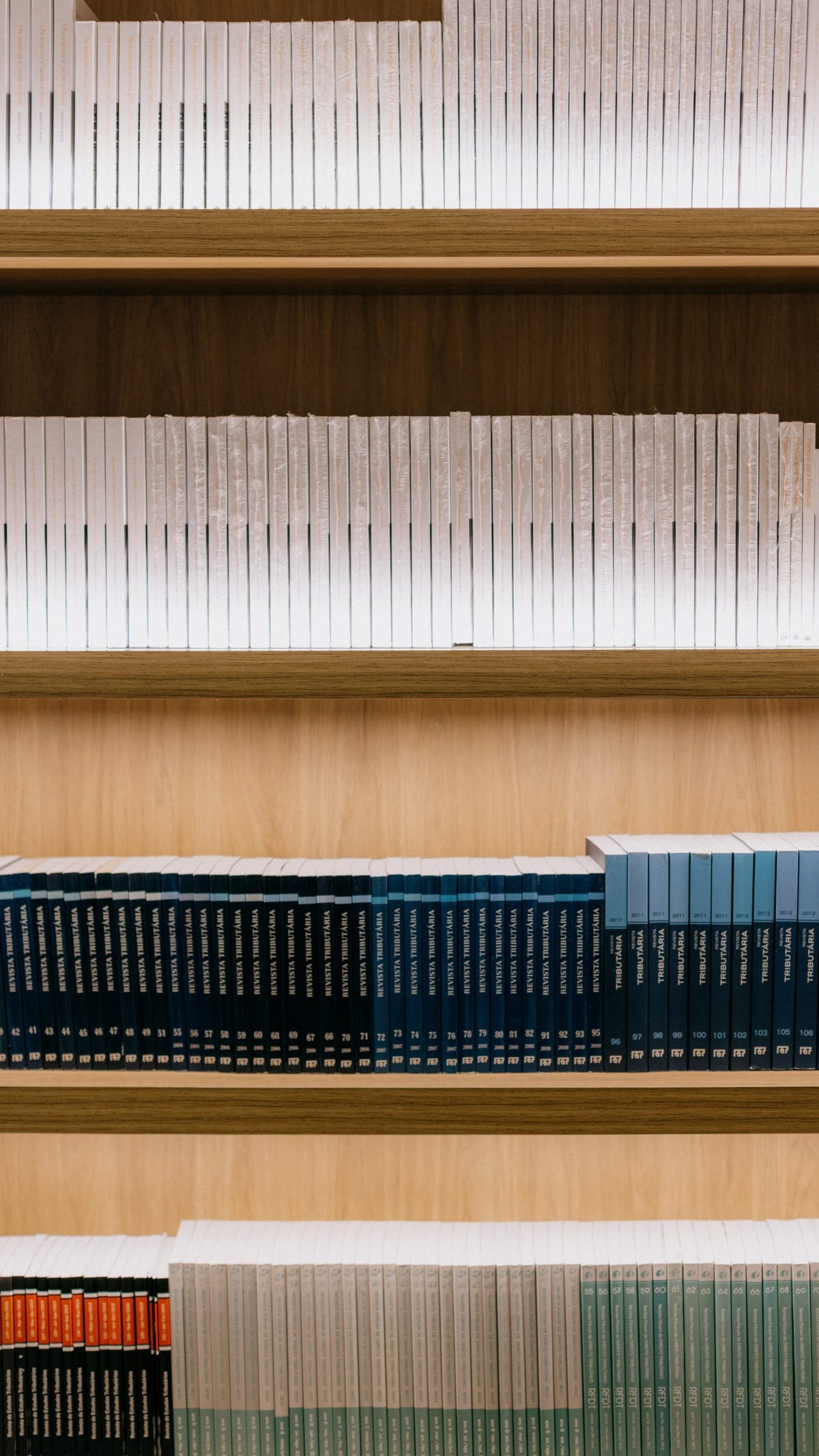 Una estantería de madera que muestra varias filas de libros uniformes, con el estante superior lleno de libros blancos, el estante medio con libros azul oscuro y turquesa, y el estante inferior que contiene una mezcla de libros claros azules y blancos, organizados cuidadosamente.