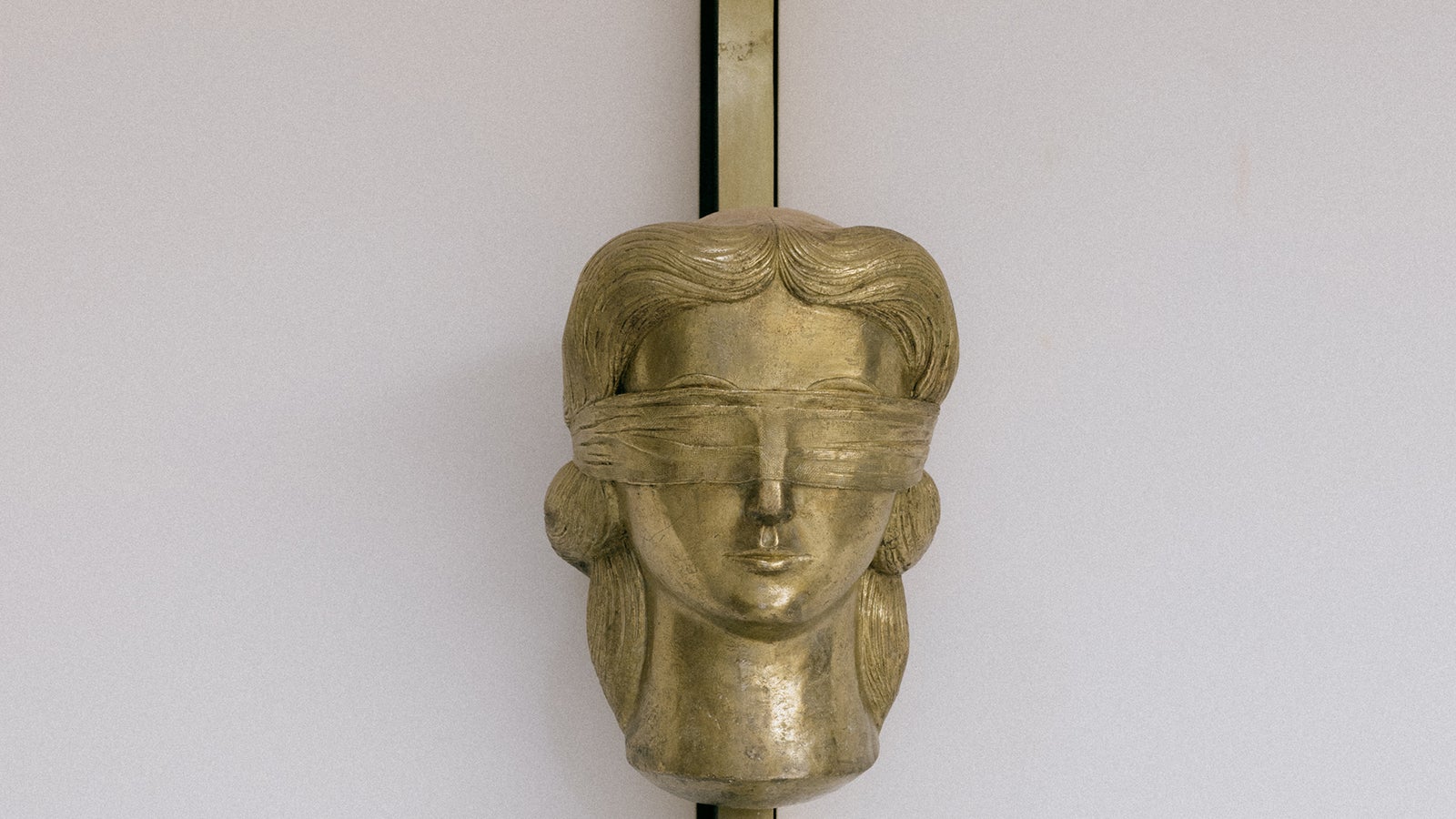 Una escultura en tono dorado de la cabeza de una figura femenina vendada con los ojos vendados, con el pelo ondulado y una cara lisa, montada en un poste vertical sobre un fondo blanco.