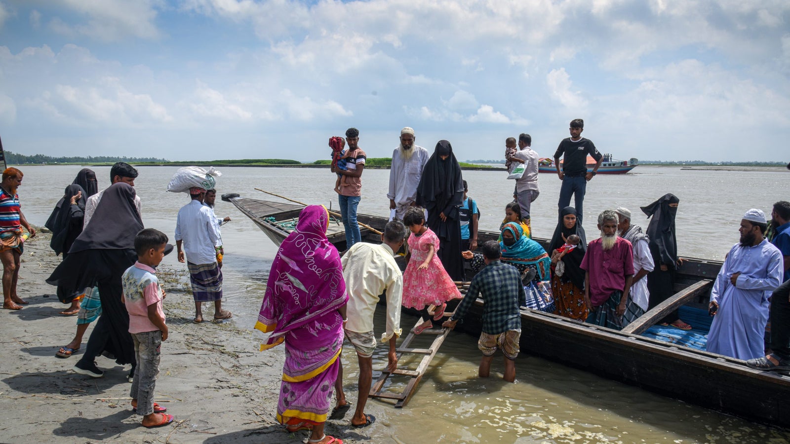 Un grupo diverso de personas se reunieron en una orilla del río preparándose para abordar un barco, con niños, hombres y mujeres con ropa tradicional. Algunos llevan bolsas y niños, mientras que otros ayudan con la carga. El fondo presenta un cielo nublado y vegetación a través del agua.